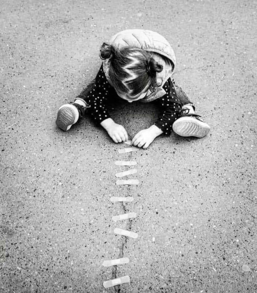 Black-and-white image of a small girl sitting on asphalt, carefully placing Band-Aids across a crack in the pavement to mimic sewing or healing.