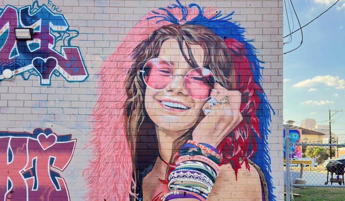 Photorealistic mural of Janis Joplin painted on a light brick wall by artist Paola Sire in San Antonio, Texas. Joplin is depicted smiling widely with pink-tinted round sunglasses, a fluffy pink and blue feathered hood, long wavy brown hair, and stacked colorful bracelets on her wrist. The mural shows her holding a hand to her cheek, radiating warmth and joy. The artist’s handle @artxsire is signed on the bottom right.