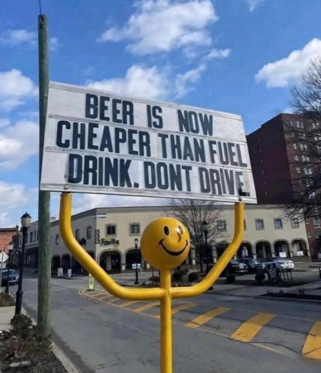 A humorous roadside sign stating 'Beer is now cheaper than fuel. Drink. Don't drive.' The sign is held by a yellow stick figure with a smiling face against a blue sky.